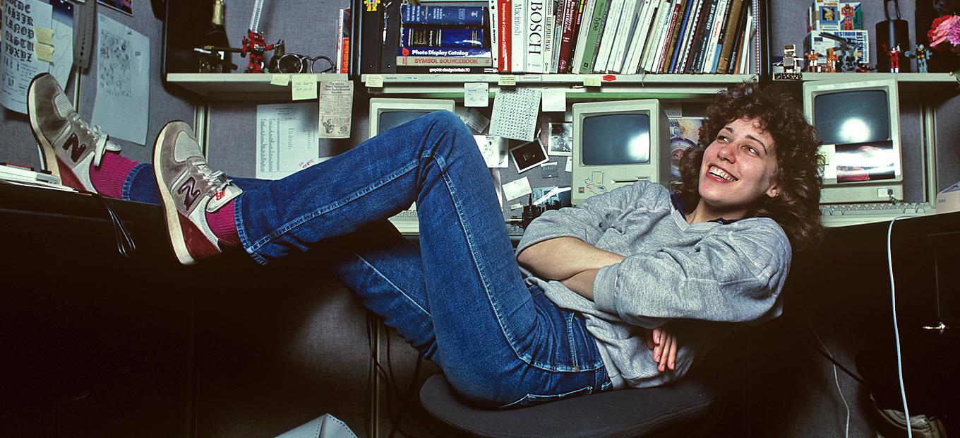 Foto von Susan Kare an ihrem Arbeitsplatz, gemütlich auf einer Couch mit Macs und Büchern im Hintergrund.